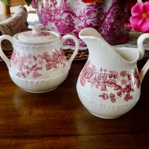 May include: A white ceramic creamer and sugar bowl set. Both pieces feature a red floral pattern with leaves and berries. The creamer has a curved spout and handle, while the sugar bowl has a lid and handles.