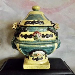May include: A decorative ceramic jar with a lid, featuring a yellow and teal color scheme. The jar is adorned with floral accents and a small basket of flowers. The jar sits on a square base.