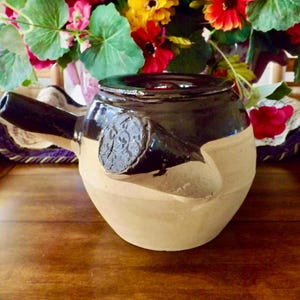 May include: A brown and beige ceramic teapot with a unique spout and a decorative design. The teapot is sitting on a wooden table.