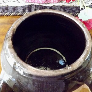 May include: A large, dark brown ceramic crock with a wide opening and two handles. The crock has a glazed finish and a white rim around the opening.