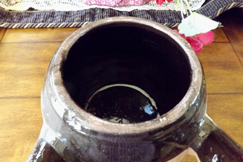 May include: A large, dark brown ceramic crock with a wide opening and two handles. The crock has a glazed finish and a white rim around the opening.