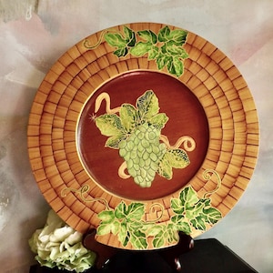 May include: Decorative plate with a basket weave border and a central maroon area. The plate features a green grape cluster and leaves design. The plate is displayed on a dark wooden stand, with a floral arrangement in the foreground.