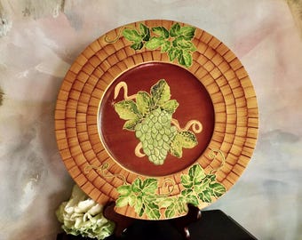 Puede incluir: Plato decorativo con borde de tejido de cesta y zona central granate. El plato presenta un diseño de racimo de uvas verdes y hojas. El plato se exhibe sobre un soporte de madera oscura, con un arreglo floral en primer plano.