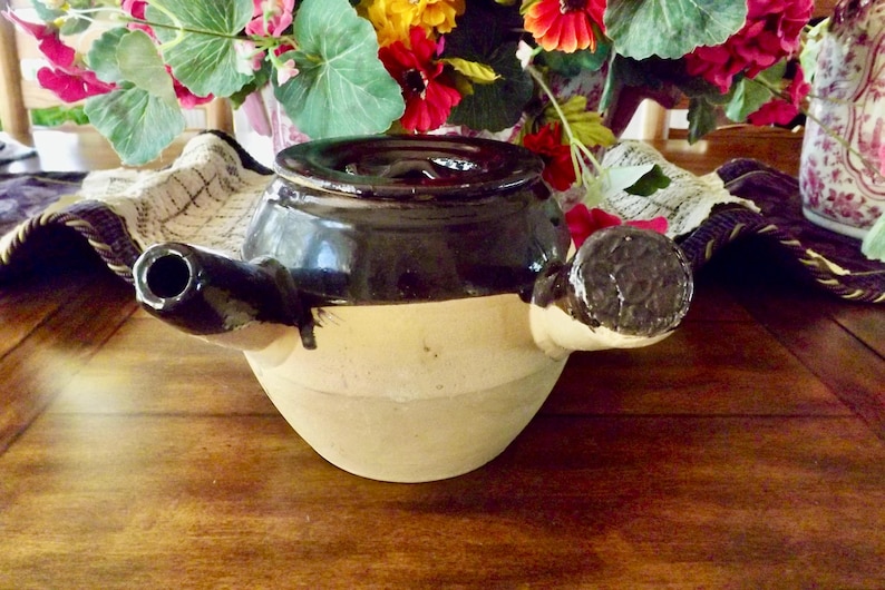 May include: A brown and beige ceramic teapot with a dark brown lid and a spout on the side. The teapot has a handle on the side and a smaller handle on the opposite side.