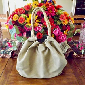 Puede incluir: Un bolso beige con cordón y asas de cuerda sobre una mesa de madera. Detrás del bolso, un vibrante ramo de flores en tonos rojos, amarillos y rosas. Dos jarrones decorativos flanquean el bolso.