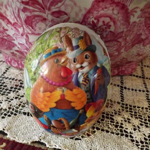 May include: A decorative Easter egg featuring a cartoon chicken and rabbit in formal attire. The egg is brightly colored with a detailed illustration, set against a floral background. The egg is resting on a lace tablecloth.