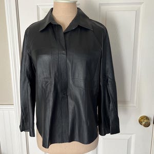 May include: A black leather button-down shirt featuring a classic collar, long sleeves with cuffs, and two rectangular chest pockets. The garment has a smooth finish, a straight hemline, and a concealed front button placket for a clean appearance.