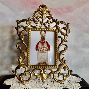 May include: Ornate gold-colored picture frame with a decorative, scrolling design. The frame holds a print of a rabbit wearing a red vest and holding a basket of Easter eggs. The print has the words "Easter Greetings".