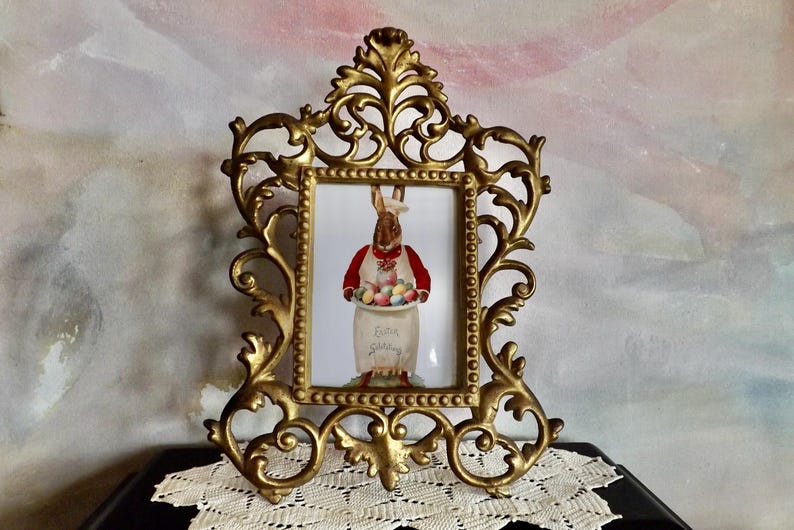 May include: Ornate gold-colored picture frame with a decorative, scrolling design. The frame holds a print of a rabbit wearing a red vest and holding a basket of Easter eggs. The print has the words "Easter Greetings".