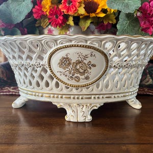 Puede incluir: Un cuenco ovalado de cer&aacute;mica con cuerpo de celos&iacute;a blanca y bordes dorados. El centro muestra un medall&oacute;n ovalado con una ilustraci&oacute;n floral marr&oacute;n. El cuenco se apoya sobre tres pies ornamentados con detalles dorados y tiene un borde festoneado.