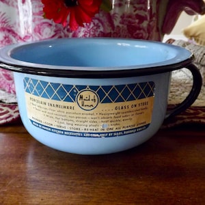 May include: A light blue porcelain enamel bowl with a black rim and handle. The bowl has a label that reads "Maid of Honor Porcelain Enamelware" and "Glass on Steel". The bowl is sitting on a wooden surface.