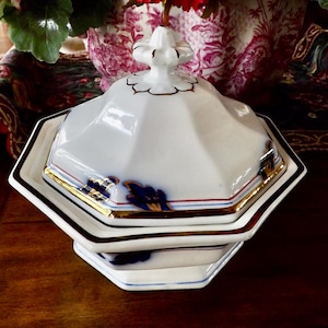May include: A white, octagonal serving dish with a lid. The dish has gold trim and blue floral accents. The lid is topped with a decorative finial. The dish is sitting on a dark wooden surface.