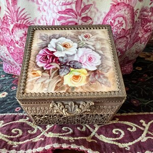 May include: A square metal jewelry box with an ornate filigree base and a hinged lid. The lid features a painted floral design of white, pink, and yellow roses on a beige background, framed by a decorative beaded gold-tone metal border.
