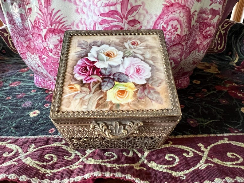 May include: A square metal jewelry box with an ornate filigree base and a hinged lid. The lid features a painted floral design of white, pink, and yellow roses on a beige background, framed by a decorative beaded gold-tone metal border.
