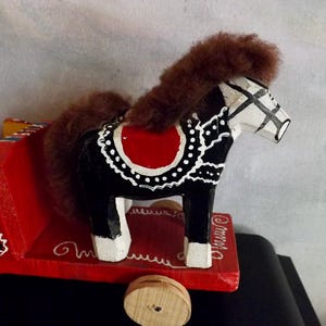Puede incluir: Un juguete de caballo de balanc&iacute;n de madera pintado de negro, blanco y rojo con una crin y cola de pelo marr&oacute;n. El caballo est&aacute; sobre una base de madera roja con ruedas.