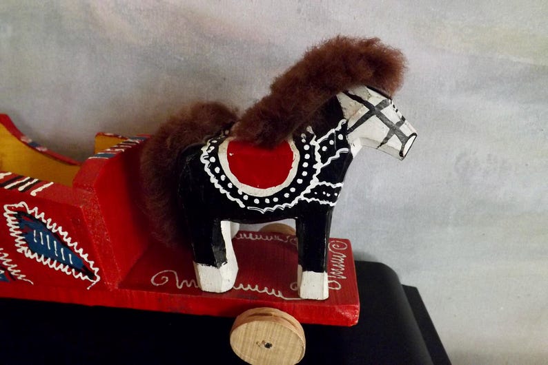 Puede incluir: Un juguete de caballo de balanc&iacute;n de madera pintado de negro, blanco y rojo con una crin y cola de pelo marr&oacute;n. El caballo est&aacute; sobre una base de madera roja con ruedas.