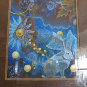 May include: A framed watercolor painting of a white rabbit with a blue and gold background. The rabbit is surrounded by leaves and orbs of light.