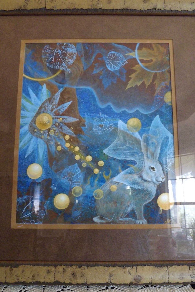 May include: A framed watercolor painting of a white rabbit with a blue and gold background. The rabbit is surrounded by leaves and orbs of light.