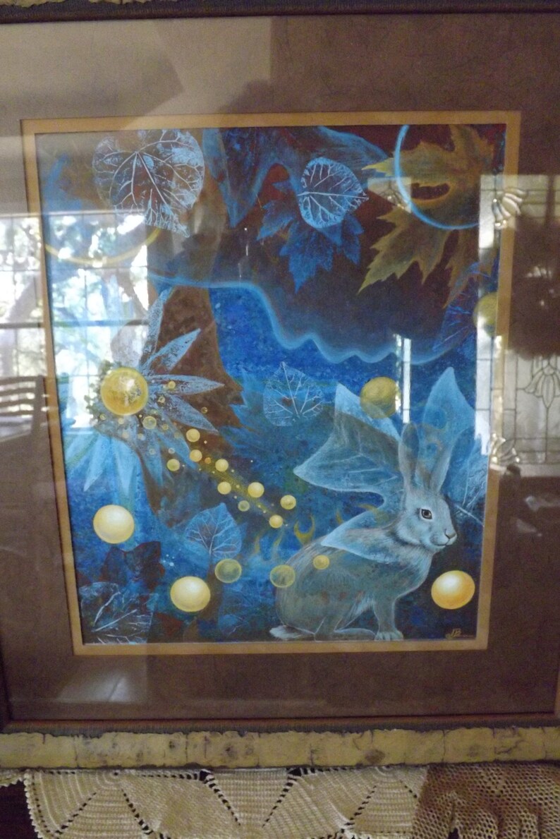 May include: A framed painting of a blue rabbit with large ears in a forest setting. The rabbit is surrounded by blue and yellow leaves and glowing orbs.