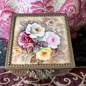 May include: A square metal trinket box featuring a decorative brass-toned frame with an ornate latch. The lid displays a painted floral arrangement of white, pink, and yellow roses set against a soft, neutral background with green leaves.