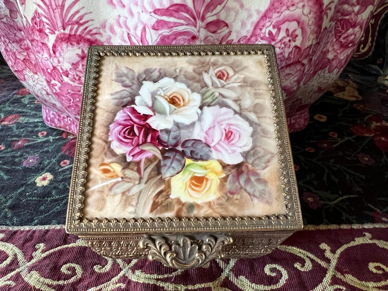 May include: A square metal trinket box featuring a decorative brass-toned frame with an ornate latch. The lid displays a painted floral arrangement of white, pink, and yellow roses set against a soft, neutral background with green leaves.
