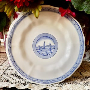May include: White ceramic plate with a blue painted border and a central design of trees and hills. The plate has a scalloped edge and is displayed on a lace tablecloth. Decorative tableware.