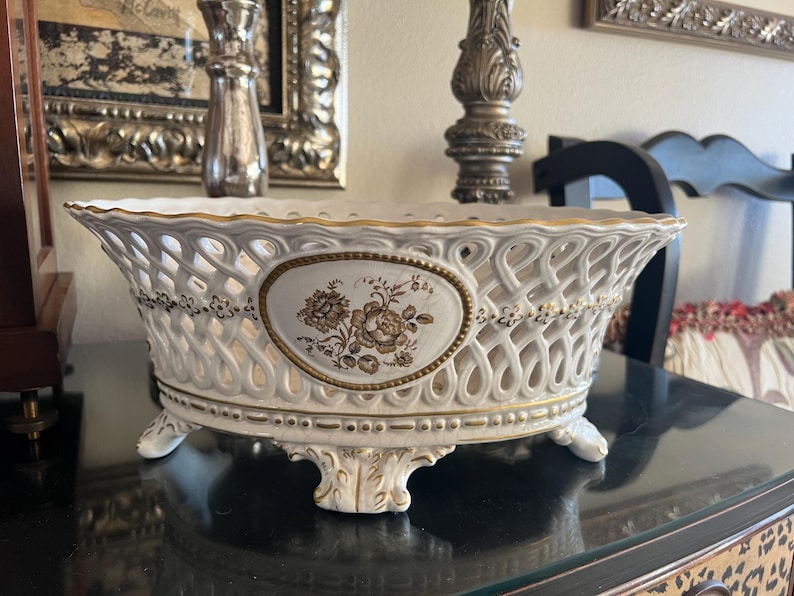Puede incluir: Un cuenco ovalado de cer&aacute;mica con un cuerpo de celos&iacute;a blanca, bordes y pies con detalles dorados. El centro del cuenco muestra un medall&oacute;n ovalado con una ilustraci&oacute;n floral marr&oacute;n detallada. El dise&ntilde;o incluye peque&ntilde;as flores doradas.