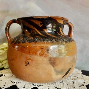 May include: A brown clay pot with two handles and a black and brown glaze. The pot has a decorative design around the top.