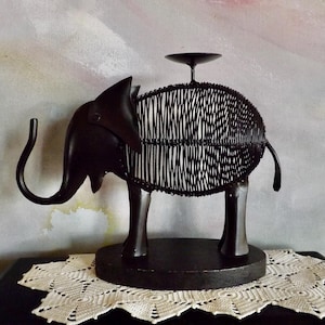 Puede incluir: Un portavelas con forma de elefante de metal negro. El elefante tiene un cuerpo tejido en forma de cesta y una pequeña plataforma circular en la espalda para una vela. El portavelas está sobre una base redonda y sobre un tapete de encaje blanco.