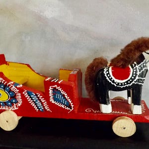 Puede incluir: Un carro de juguete de madera rojo con un caballo blanco y negro tirando de &eacute;l. El carro est&aacute; decorado con flores y remolinos coloridos. El caballo tiene una melena y una cola marrones.