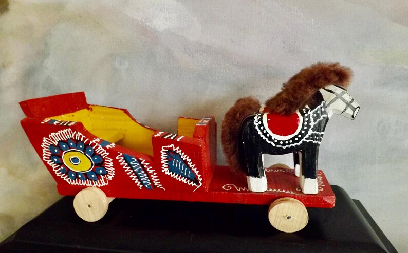 Puede incluir: Un carro de juguete de madera rojo con un caballo blanco y negro tirando de &eacute;l. El carro est&aacute; decorado con flores y remolinos coloridos. El caballo tiene una melena y una cola marrones.