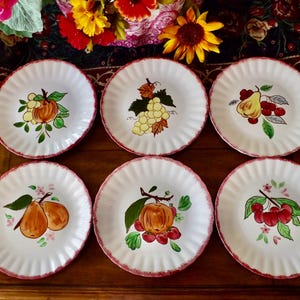 May include: Six white ceramic plates with scalloped edges and a red trim. Each plate features a different hand-painted fruit design, including pumpkins, grapes, pears, and cherries. The plates are arranged on a wooden surface.