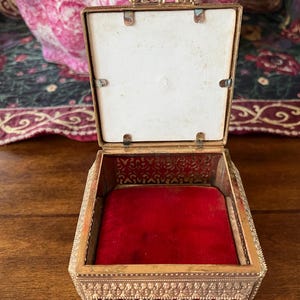 May include: A square metal trinket box with an ornate gold-tone filigree exterior and a hinged lid. The interior is lined with a vibrant red velvet base and features a white square panel on the inside of the lid, secured by four small metal tabs.