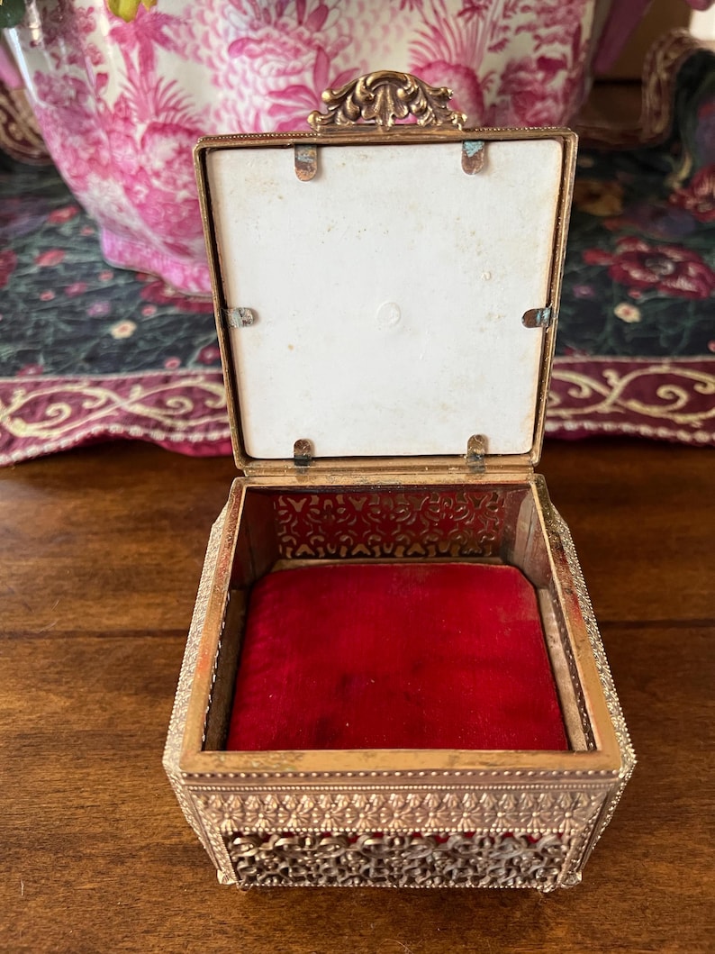May include: A square metal trinket box with an ornate gold-tone filigree exterior and a hinged lid. The interior is lined with a vibrant red velvet base and features a white square panel on the inside of the lid, secured by four small metal tabs.