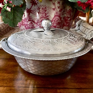 May include: A silver-colored, lidded serving dish with floral and leaf patterns. The dish has a hammered texture on the bowl and a decorative handle on the lid. The dish is on a wooden table, with flowers and a patterned cloth in the background.