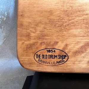 May include: A light brown wooden board featuring a black oval stamp. The stamp contains the text 1854, THE OLD DRUM SHOP, and GRANVILLE, MASS. The wood grain is visible across the surface, and the board has rounded corners on the right side.