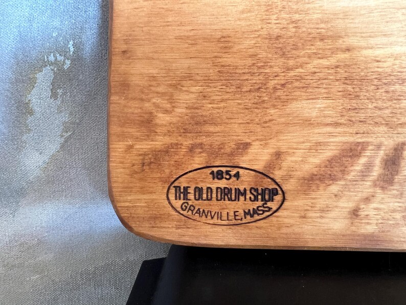 May include: A light brown wooden board featuring a black oval stamp. The stamp contains the text 1854, THE OLD DRUM SHOP, and GRANVILLE, MASS. The wood grain is visible across the surface, and the board has rounded corners on the right side.