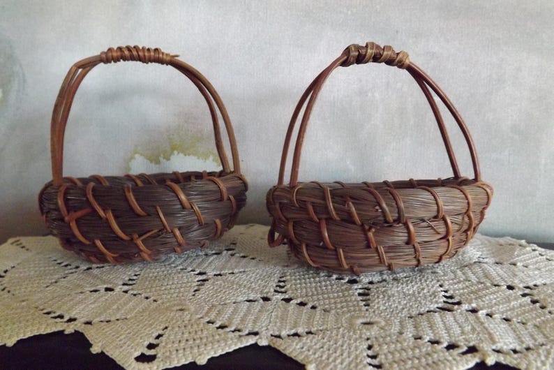 Puede incluir: Dos peque&ntilde;as cestas tejidas marrones hechas a mano con asas arqueadas. Las cestas est&aacute;n hechas de materiales naturales y se asientan sobre un mantel de encaje blanco. Las asas est&aacute;n envueltas para un agarre seguro.