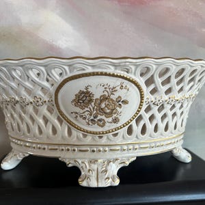 Puede incluir: Un cuenco ovalado de cer&aacute;mica con cuerpo de celos&iacute;a blanca y bordes pintados en oro. El centro muestra un medall&oacute;n ovalado con una ilustraci&oacute;n floral marr&oacute;n. El cuenco se apoya sobre cuatro patas ornamentadas y tiene un borde festoneado.