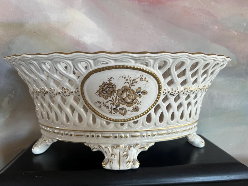 Puede incluir: Un cuenco ovalado de cer&aacute;mica con cuerpo de celos&iacute;a blanca y bordes pintados en oro. El centro muestra un medall&oacute;n ovalado con una ilustraci&oacute;n floral marr&oacute;n. El cuenco se apoya sobre cuatro patas ornamentadas y tiene un borde festoneado.