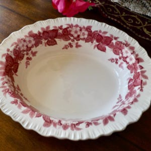 May include: Oval serving dish with a white base and a decorative floral pattern in shades of red. The rim has a scalloped edge. The dish is on a dark wooden surface.