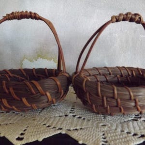 Puede incluir: Dos peque&ntilde;as cestas tejidas marrones hechas a mano con asas arqueadas. Las cestas est&aacute;n hechas de materiales naturales, con una base redonda y un dise&ntilde;o tejido. Est&aacute;n sobre una superficie de encaje blanco.