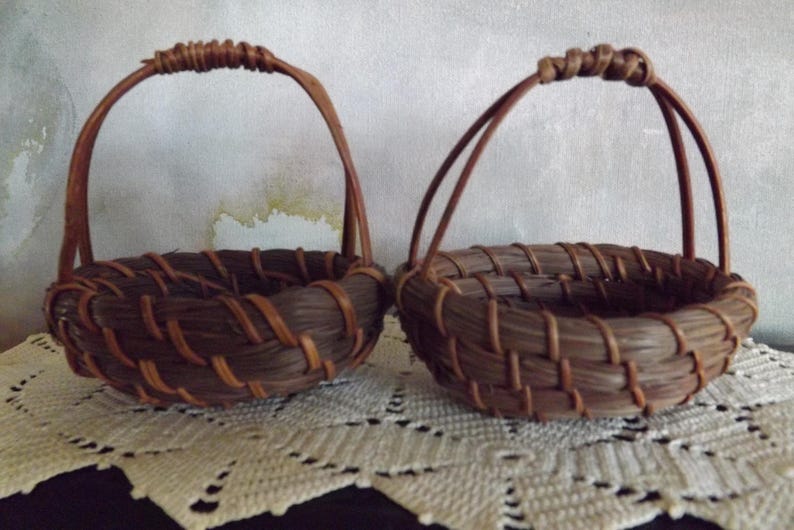 Puede incluir: Dos peque&ntilde;as cestas tejidas marrones hechas a mano con asas arqueadas. Las cestas est&aacute;n hechas de materiales naturales, con una base redonda y un dise&ntilde;o tejido. Est&aacute;n sobre una superficie de encaje blanco.