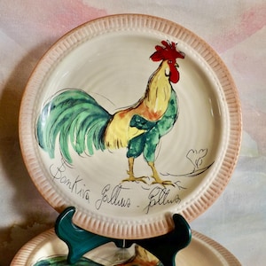 May include: A decorative ceramic plate featuring a hand-painted rooster design. The rooster has a red comb, yellow body, and green tail feathers. The plate has a cream-colored center and a pink, ridged border. The plate is supported by a dark green stand.