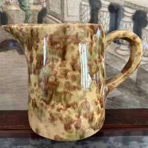 May include: A ceramic pitcher featuring a mottled glaze in shades of light green, tan, and dark brown. The vessel has a rounded body, a curved handle on the right side, and a distinct pouring spout on the left, with a glossy, smooth finish throughout.