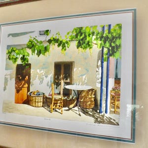 May include: A watercolor painting of a patio scene with a table and chairs, a vine growing over the wall, and baskets of fruit. The painting is framed in a light brown wood frame.
