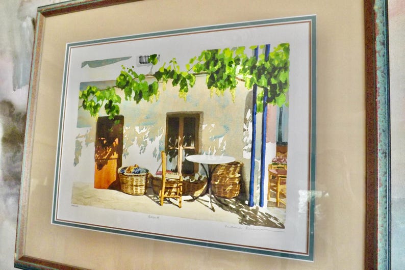May include: A watercolor painting of a patio scene with a table and chairs, a vine growing over the wall, and baskets of fruit. The painting is framed in a light brown wood frame.
