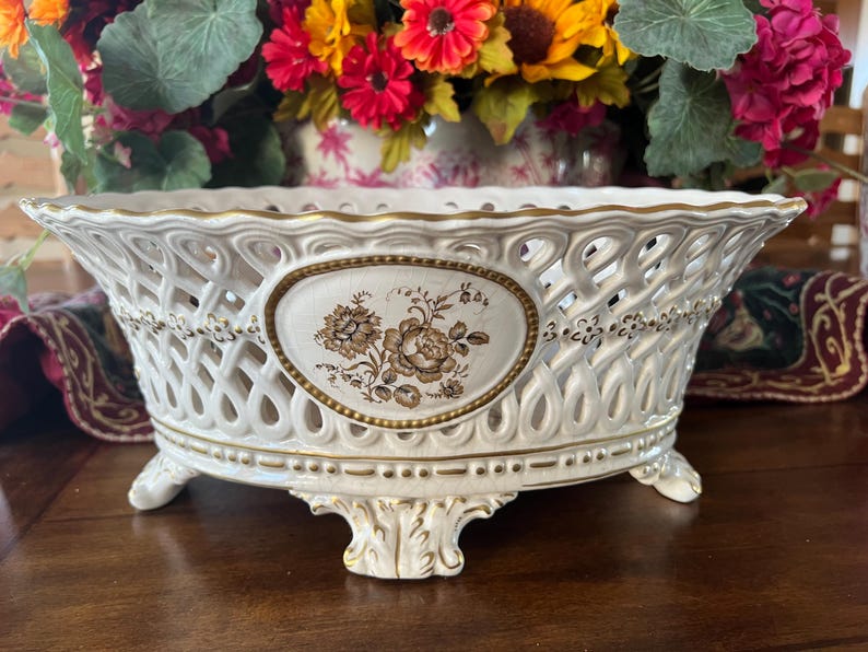 Puede incluir: Un cuenco ovalado de cer&aacute;mica con un dise&ntilde;o lateral de celos&iacute;a intrincado y un medall&oacute;n central con un estampado floral marr&oacute;n. El cuenco tiene un esmalte de color crema con detalles en oro y se apoya sobre cuatro pies esculpidos.