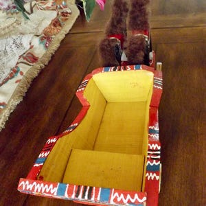 Puede incluir: Un carro de juguete de madera con dos caballos de peluche marrones tirando de &eacute;l. El carro est&aacute; pintado de rojo con rayas amarillas, azules y blancas.