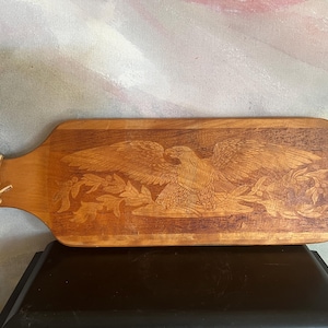 May include: A rectangular wooden serving board with a handle featuring a hole and a leather tie. The surface displays a detailed laser-engraved image of a bald eagle with wings spread wide, perched above a branch with leaves and berries.